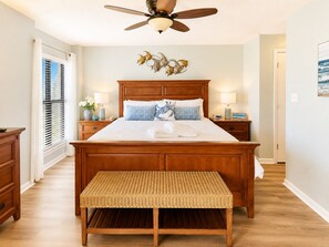 A bedroom with a large bed, wooden headboard, bedside lamps, a ceiling fan, and a wall-mounted artwork. - Summer Winds B-338, Oceanfront Condo in Indian Beach, NC! (Indian Beach)