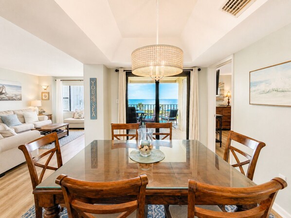 A dining area with a glass table and wooden chairs, a chandelier, and a view of the sea through an open door.
