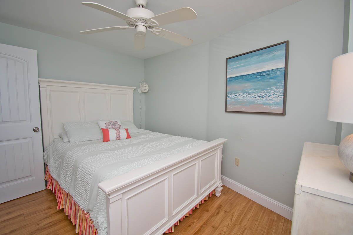 Une chambre avec un lit blanc, un ventilateur de plafond, un tableau représentant une plage et une commode blanche.