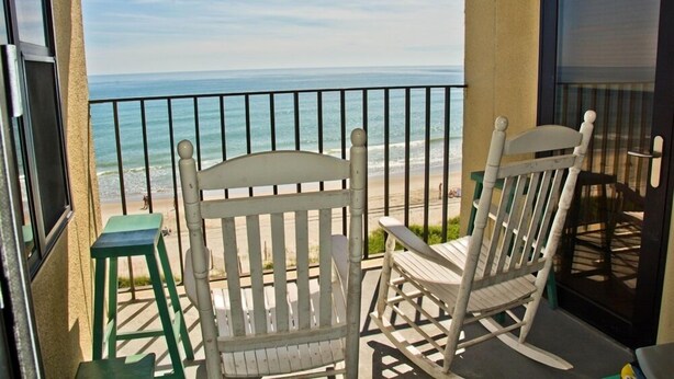 Un balcon avec deux fauteuils à bascule et un petit tabouret, donnant sur une plage avec l’océan en toile de fond.