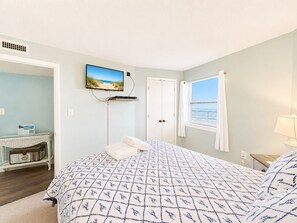 A bedroom with a bed, a television mounted on the wall, a window with curtains, and a bedside table with a lamp. - Summer Winds B-439, Oceanfront Condo in Indian Beach, NC! (Indian Beach)