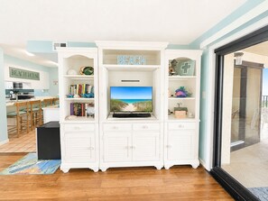 A living room with a television, bookshelves, and a kitchen area in the background. - Summer Winds B-439, Oceanfront Condo in Indian Beach, NC! (Indian Beach)