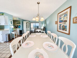 A dining area with a long table, white chairs, and a chandelier. - Summer Winds B-439, Oceanfront Condo in Indian Beach, NC! (Indian Beach)