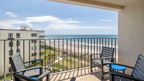 Un balcón con vista a la playa, una silla, una mesa y un edificio al fondo.