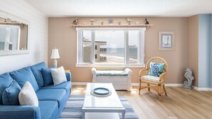 Una sala de estar con un sofá azul, un sillón de mimbre, una mesa de centro blanca y vista a la playa a través de amplios ventanales.