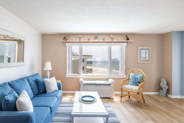 A living room with a blue sofa, a wicker chair, a white coffee table, and a view of the beach through large windows.