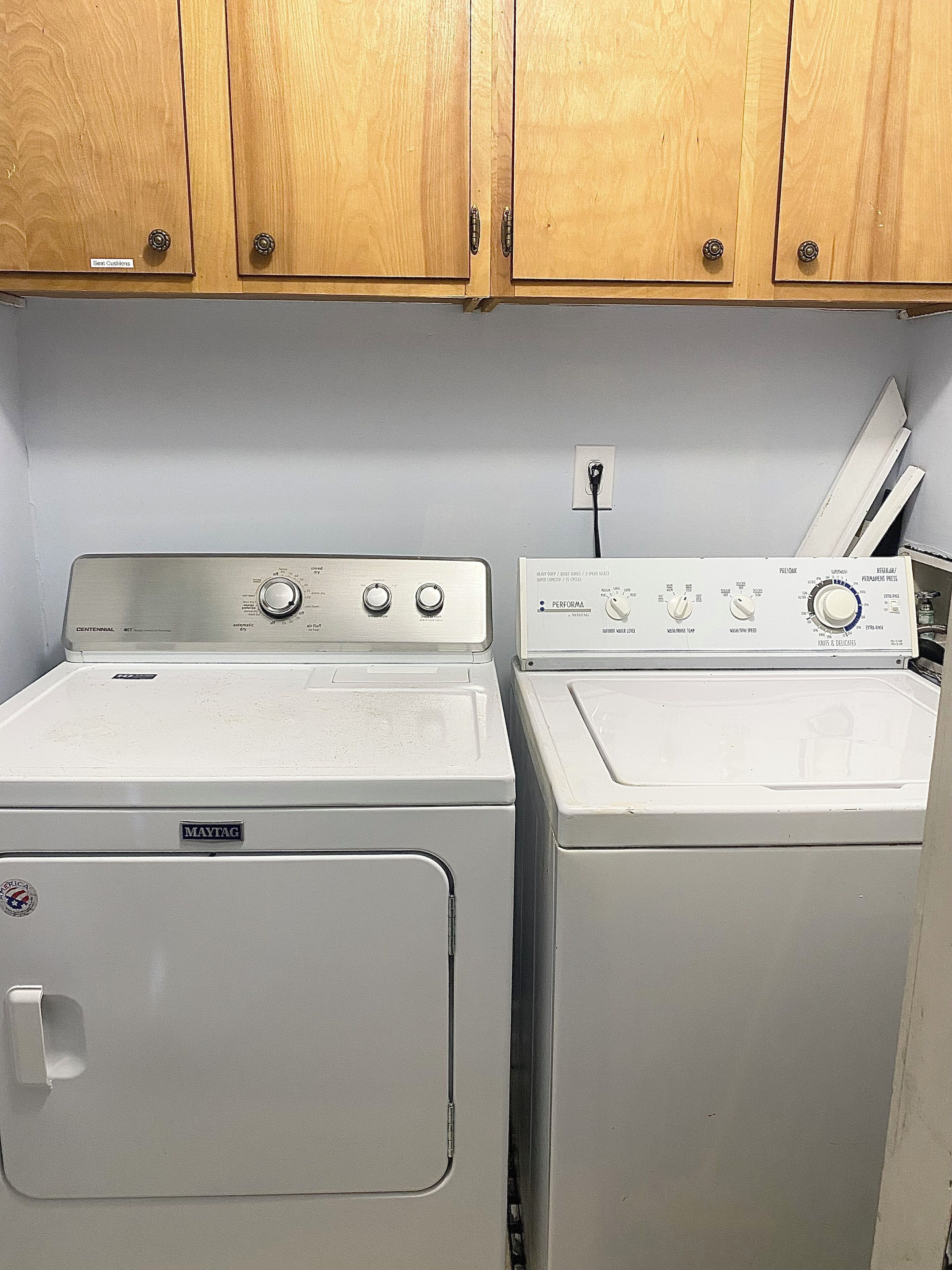 A laundry room with two washing machines and a dryer, wooden cabinets above, and a power outlet on the wall.