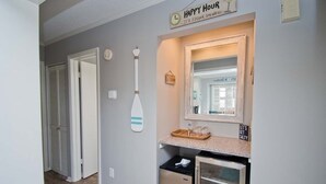 Un pasillo con una zona de cocina, que incluye un refrigerador, un microondas y un lavamanos. Hay relojes en la pared y un letrero que dice “HAPPY HOUR”.