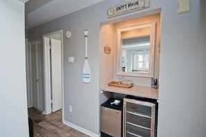 A hallway with a kitchen area, including a refrigerator, a microwave, and a sink. There are clocks on the wall and a sign that reads "HAPPY HOUR."