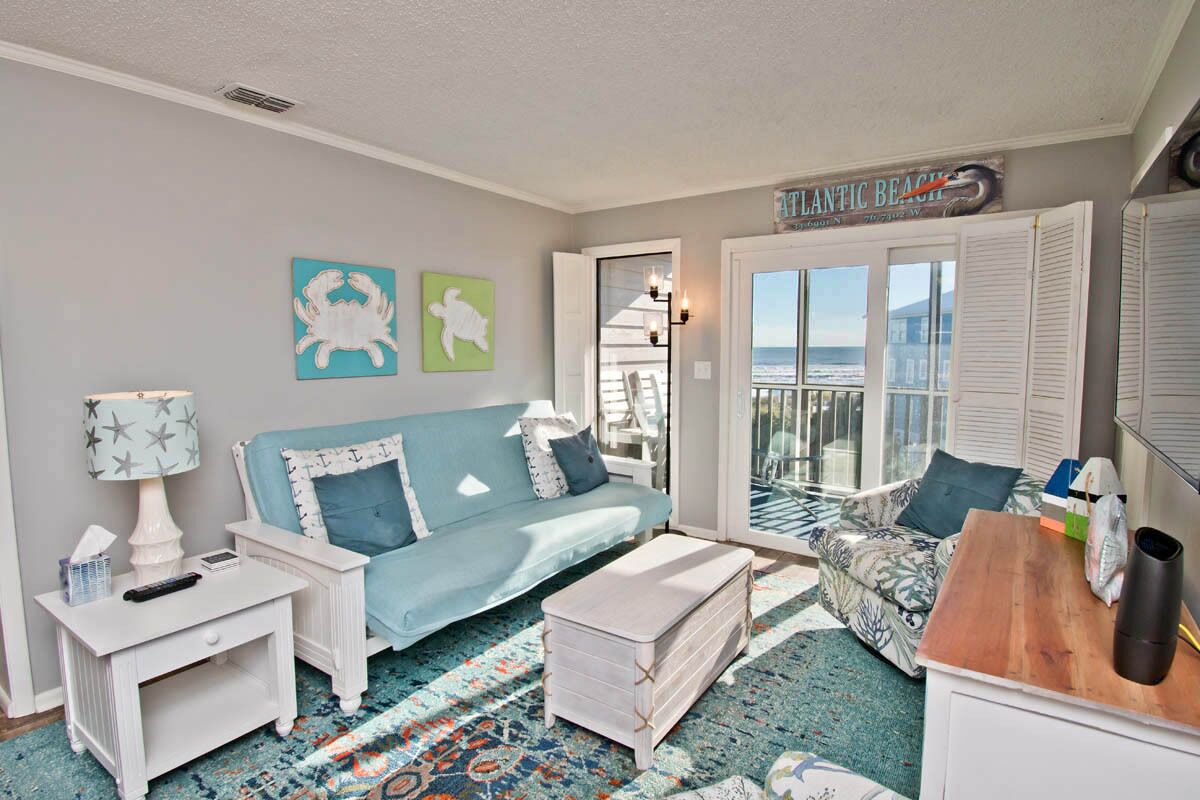 A cozy living room with a blue sofa, white side tables, and a patterned rug. A wooden coffee table and a small white cabinet are also visible.