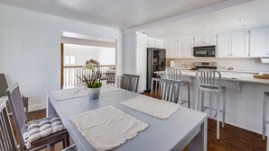 A modern kitchen with white cabinets, a microwave, and a refrigerator. There is a dining area with a table and chairs, and a potted plant on the table. - Your Spring & Summer Coastal Retreat Awaits in Huntington Beach!  (Huntington Beach)