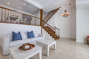 Un salon avec un escalier, un canapé blanc agrémenté de coussins bleus, une table basse blanche et un lustre.