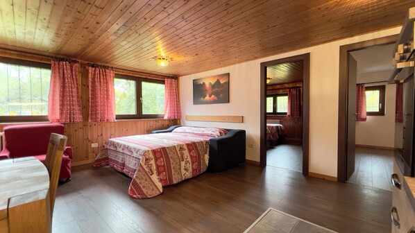 A wooden-paneled room with a bed, a chair, and a small table. There are windows with curtains, a painting on the wall, and a door leading to another room. - Les Crouses Taverne Apartments (Valtournenche)