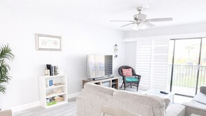 A modern living room with a sofa, a TV, a ceiling fan, and a balcony with white shutters. - Ocean view 2 bedroom townhouse (ST. AUGUSTINE)