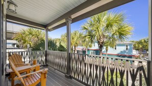 Une terrasse en bois avec des fauteuils Adirondack, une balustrade et une vue sur une maison bleue bordée de palmiers.