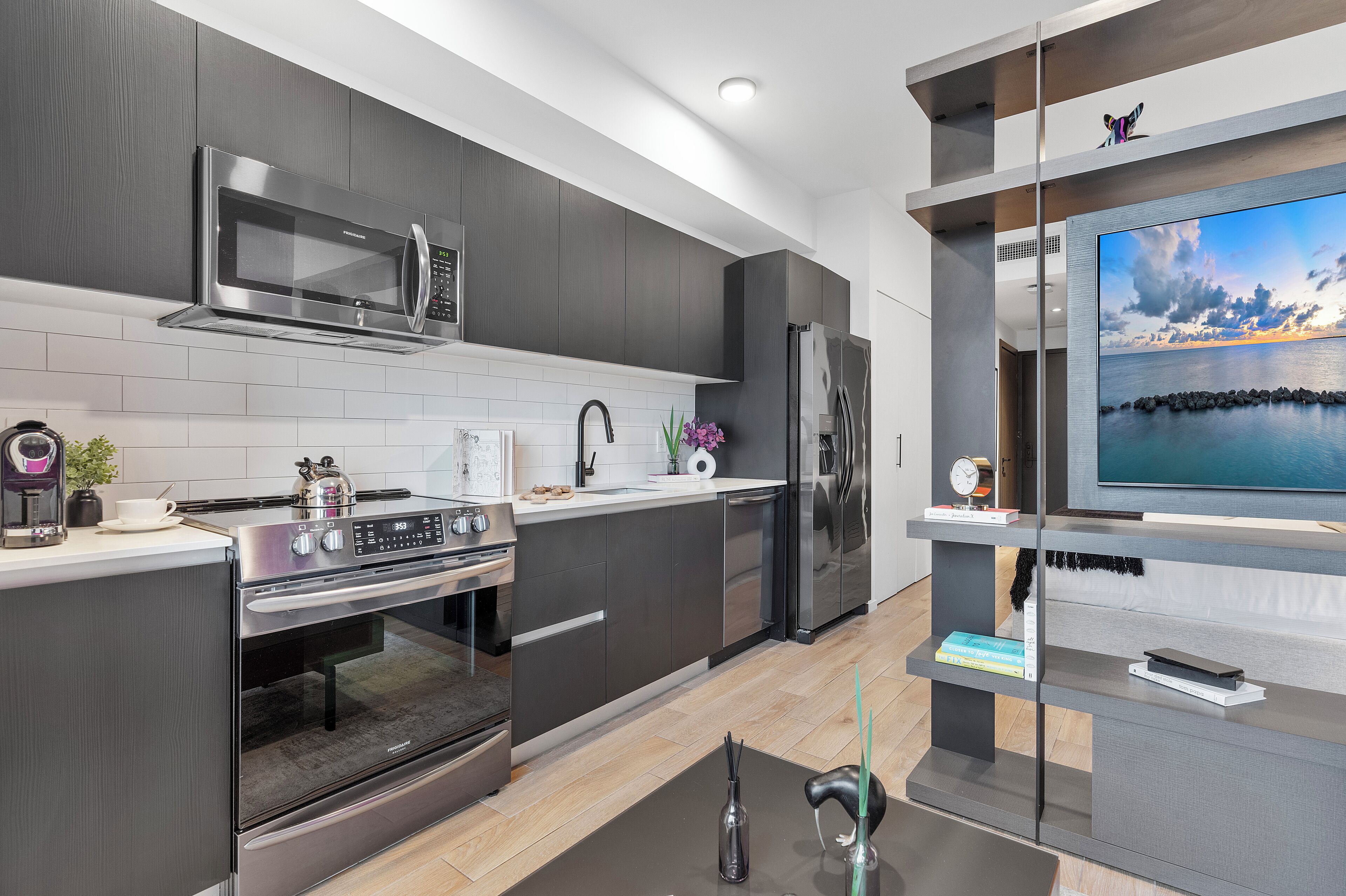 Une cuisine moderne avec des meubles de couleur sombre, des appareils en acier inoxydable et un téléviseur à écran plat posé sur une étagère ouverte.