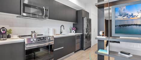 Une cuisine moderne avec des meubles de couleur sombre, des appareils en acier inoxydable et un téléviseur à écran plat posé sur une étagère ouverte.
