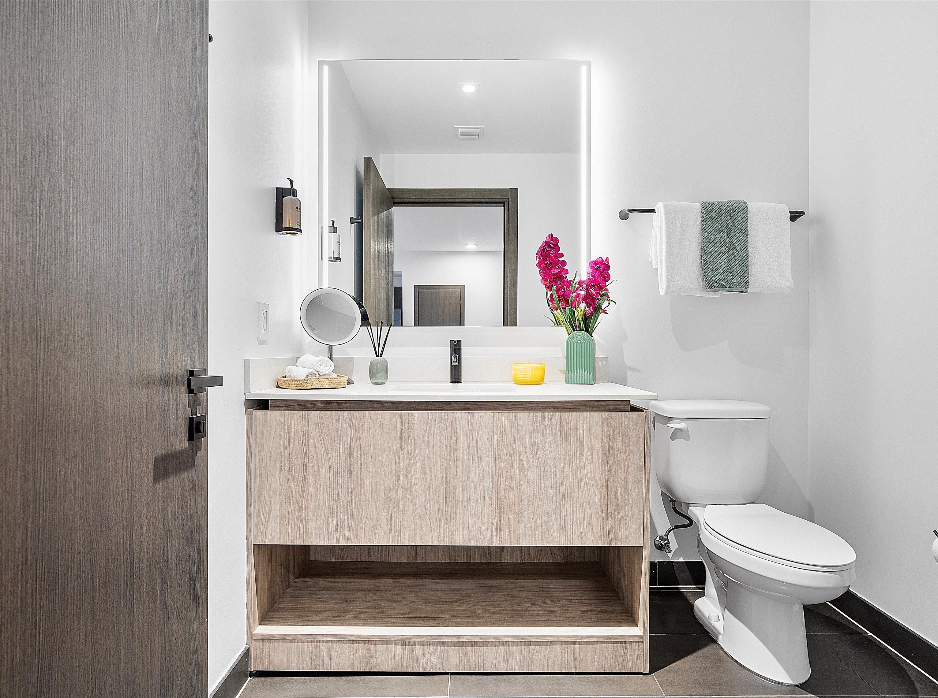 Une salle de bain moderne avec un meuble-lavabo en bois, un miroir, des toilettes et un vase rempli de fleurs.