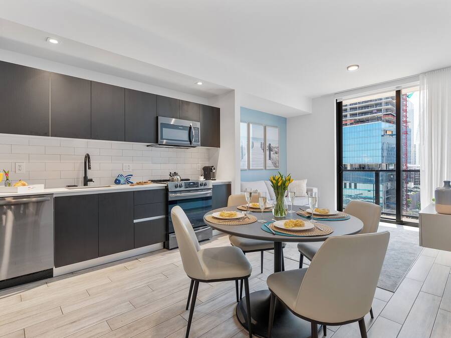 A modern kitchen with dark cabinets, a stainless steel dishwasher, and a microwave. There is a dining area with a round table, chairs, and a vase of flowers.
