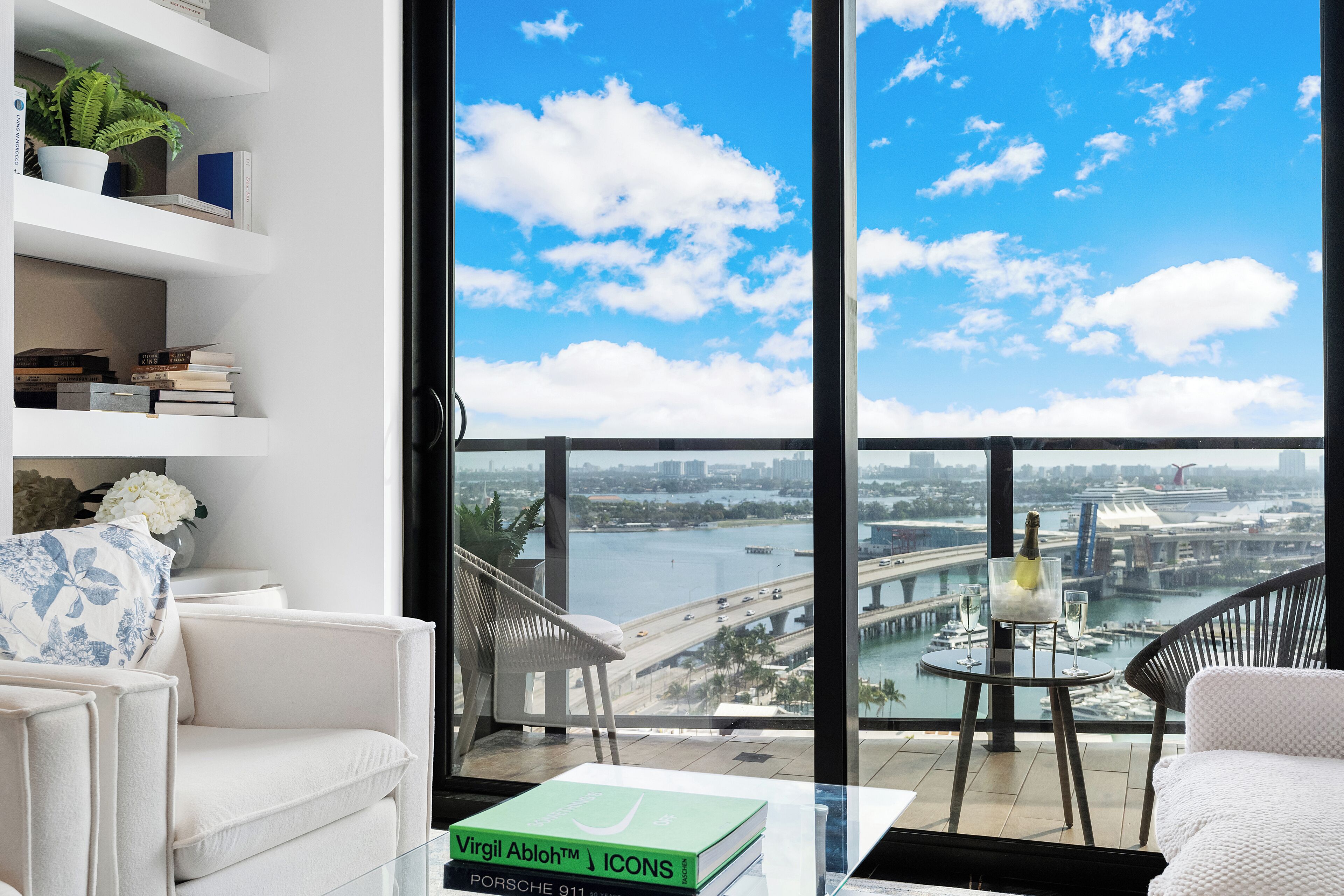 A modern living room with a view of a city skyline, a balcony with a table and chairs, and a bookshelf with books and decorative items.