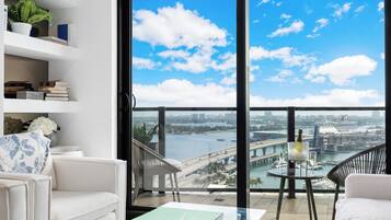 A modern living room with a view of a city skyline, a balcony with a table and chairs, and a bookshelf with books and decorative items.