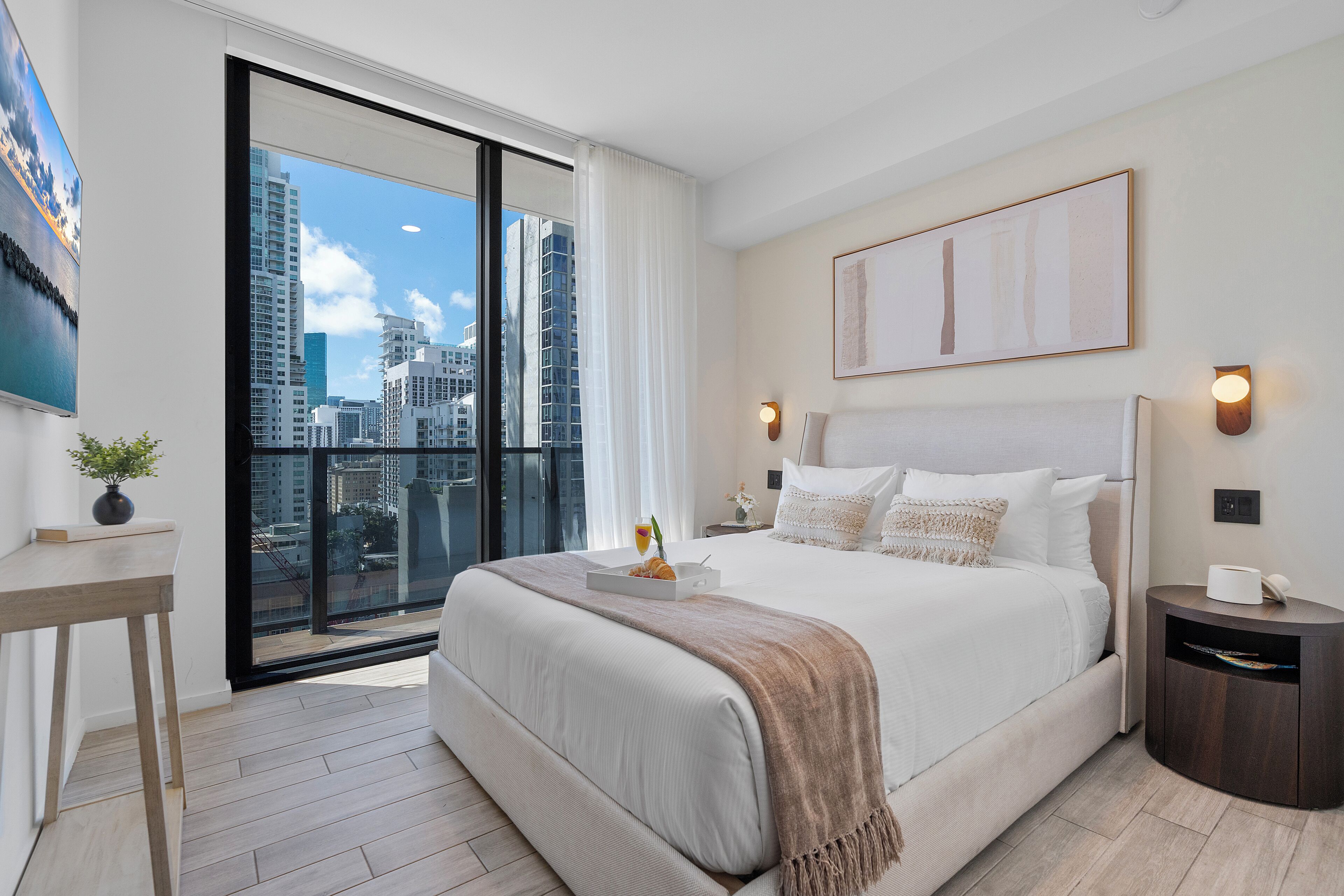 A modern hotel room with a large bed, a bedside table, a nightstand, a city view through a large window, and a painting on the wall.