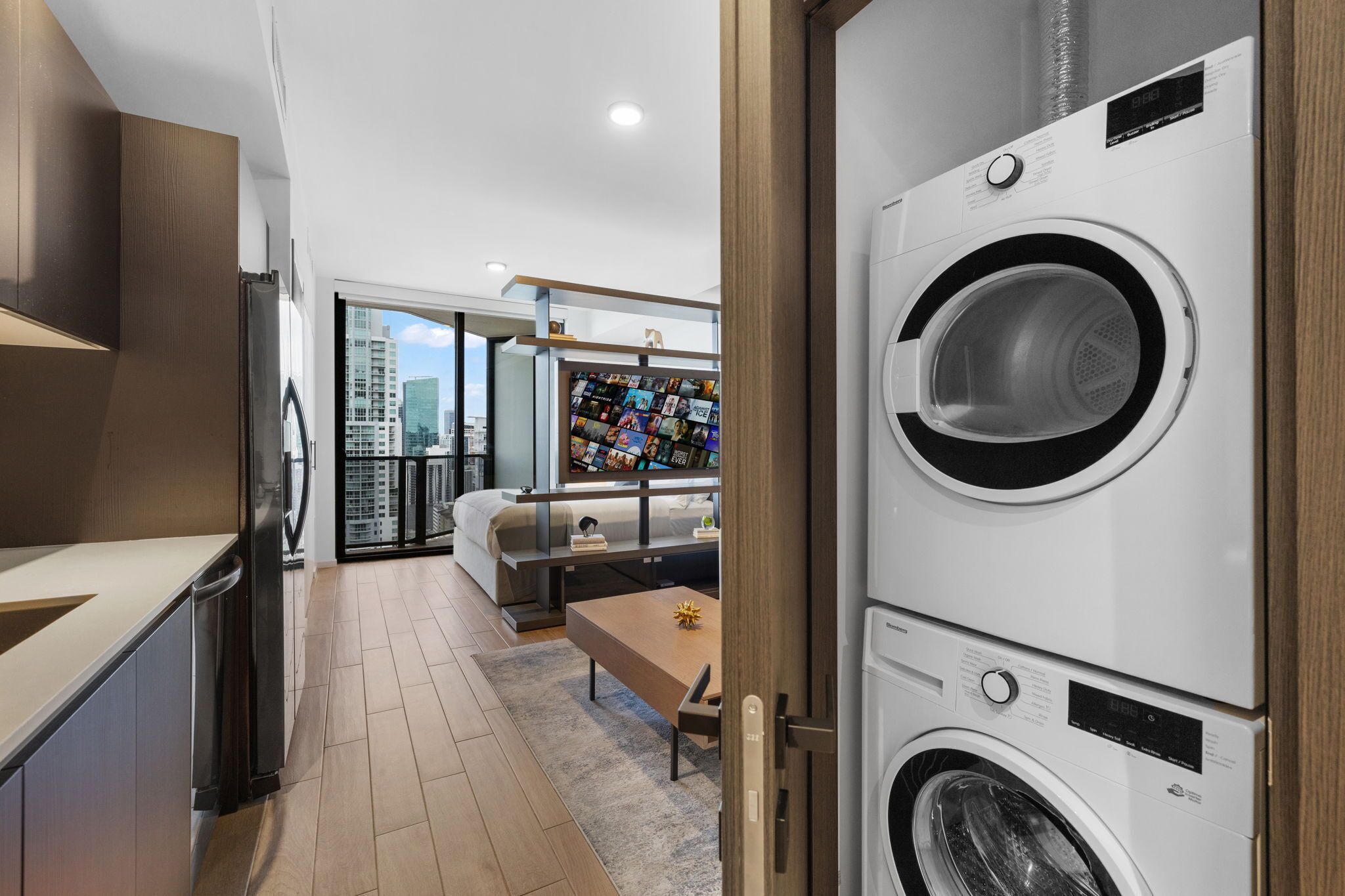 A modern laundry room with stacked washers and dryers, a dining area with a bench, and a view of the city through large windows.