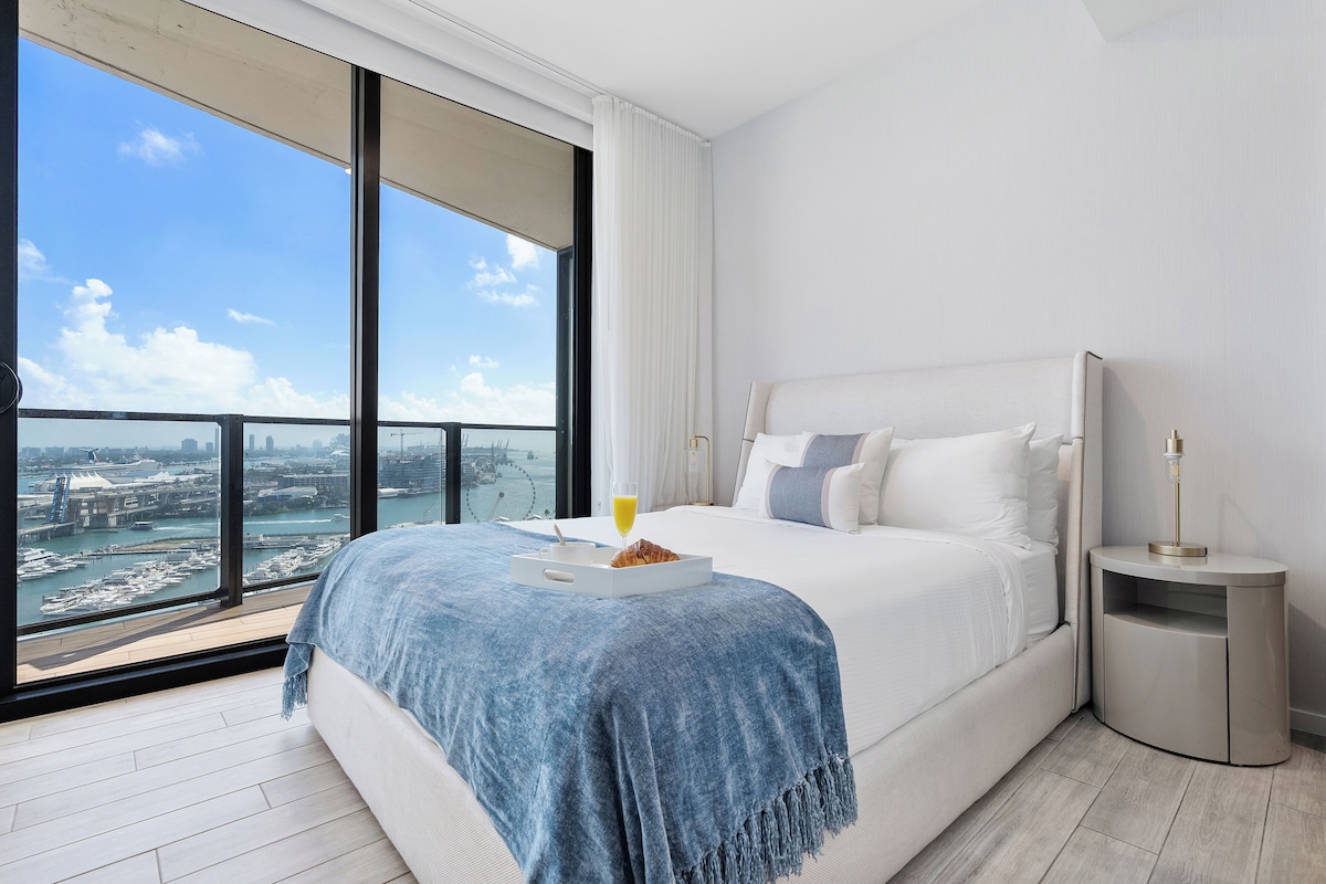A modern hotel room with a large bed, a bedside table, a nightstand, and a view of a cityscape through large windows.
