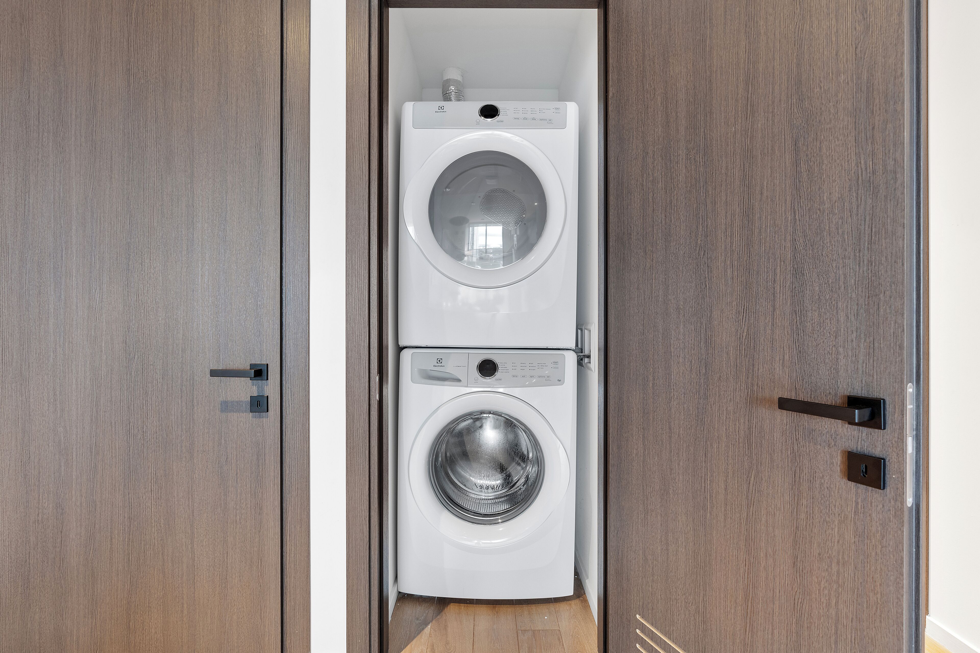 A compact, modern laundry room with a built-in washer and dryer, featuring a wooden door and a metal handle.