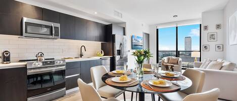 A modern kitchen with a built-in microwave, oven, and refrigerator. There is a dining area with a round table and chairs, and a view of a cityscape through large windows.
