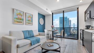 A modern living room with a sofa, coffee table, and a dining area with a view of the cityscape through large windows.