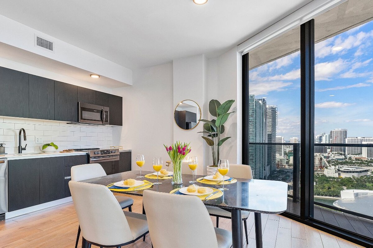 A modern kitchen with dark cabinets and a large window offering an oceanfront view. The dining area has a table set for four with yellow placemats and orange drinks. Text indicates it's on Floor 26 with no fees.