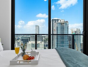 A hotel room with a large window offering a city view, a bed, a tray with croissants, strawberries, a glass of orange juice, and a cup. - Miami Vacation Rentals - Downtown (Miami)