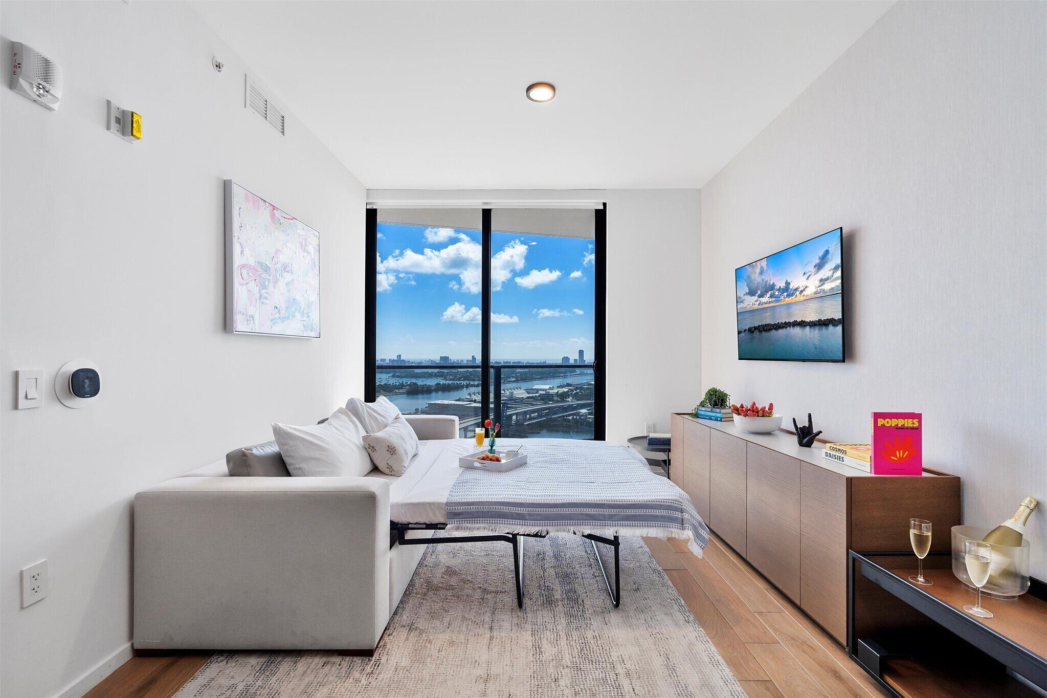 A modern hotel room with a large window, a sofa, a bedside table with a bottle and glasses, and a view of the cityscape.