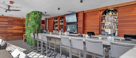 A bar area with a long counter, bar stools, a television screen, and a ceiling fan.