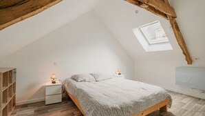 Een slaapkamer met een schuine kap, dakraam, bed met een geborduurd dekbed, nachtkastjes met lampen en een houten garderobe.
