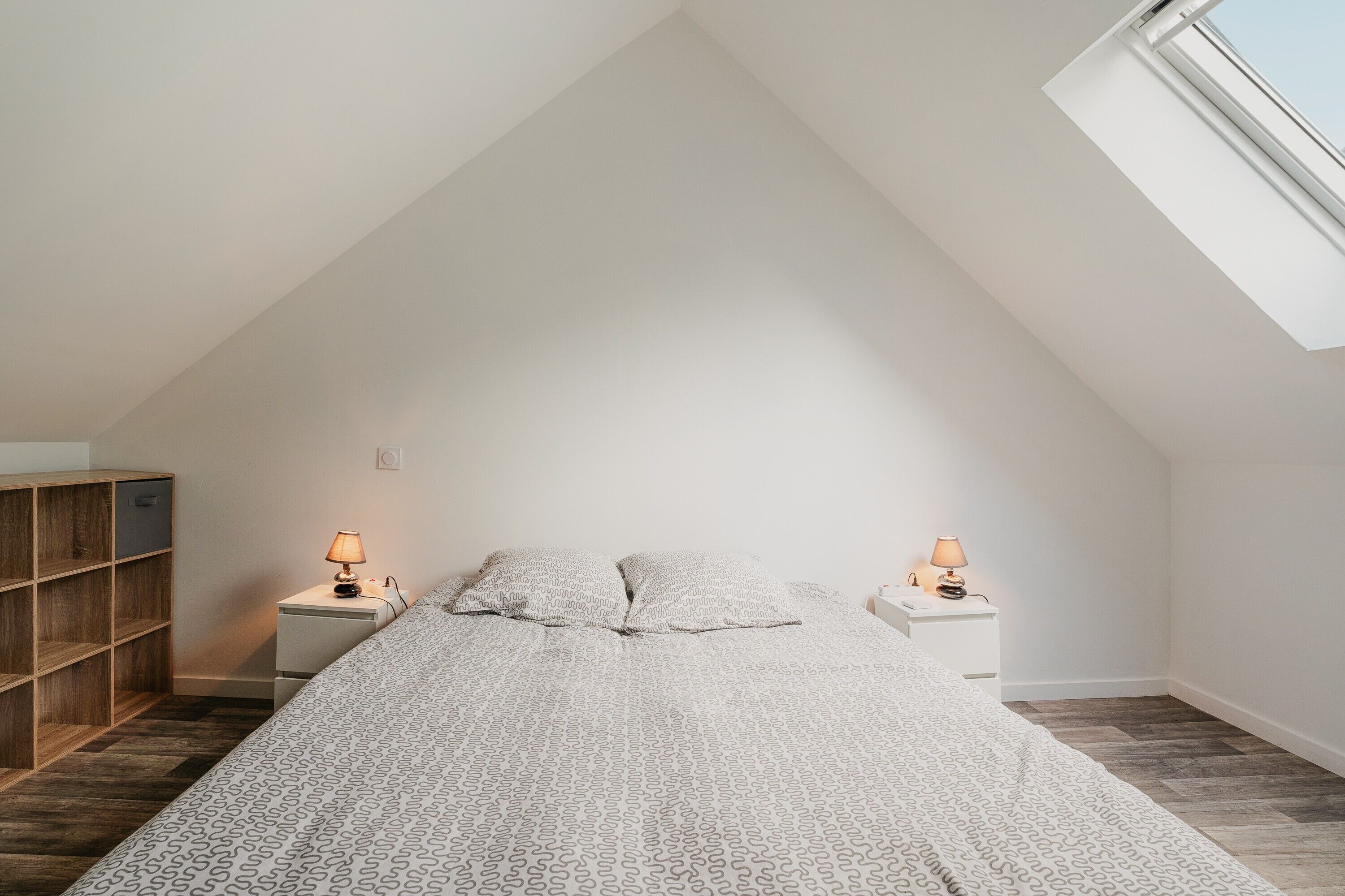 A bedroom with a sloped ceiling, a bed with patterned bedding, two bedside lamps, a wooden bookshelf, and a window.