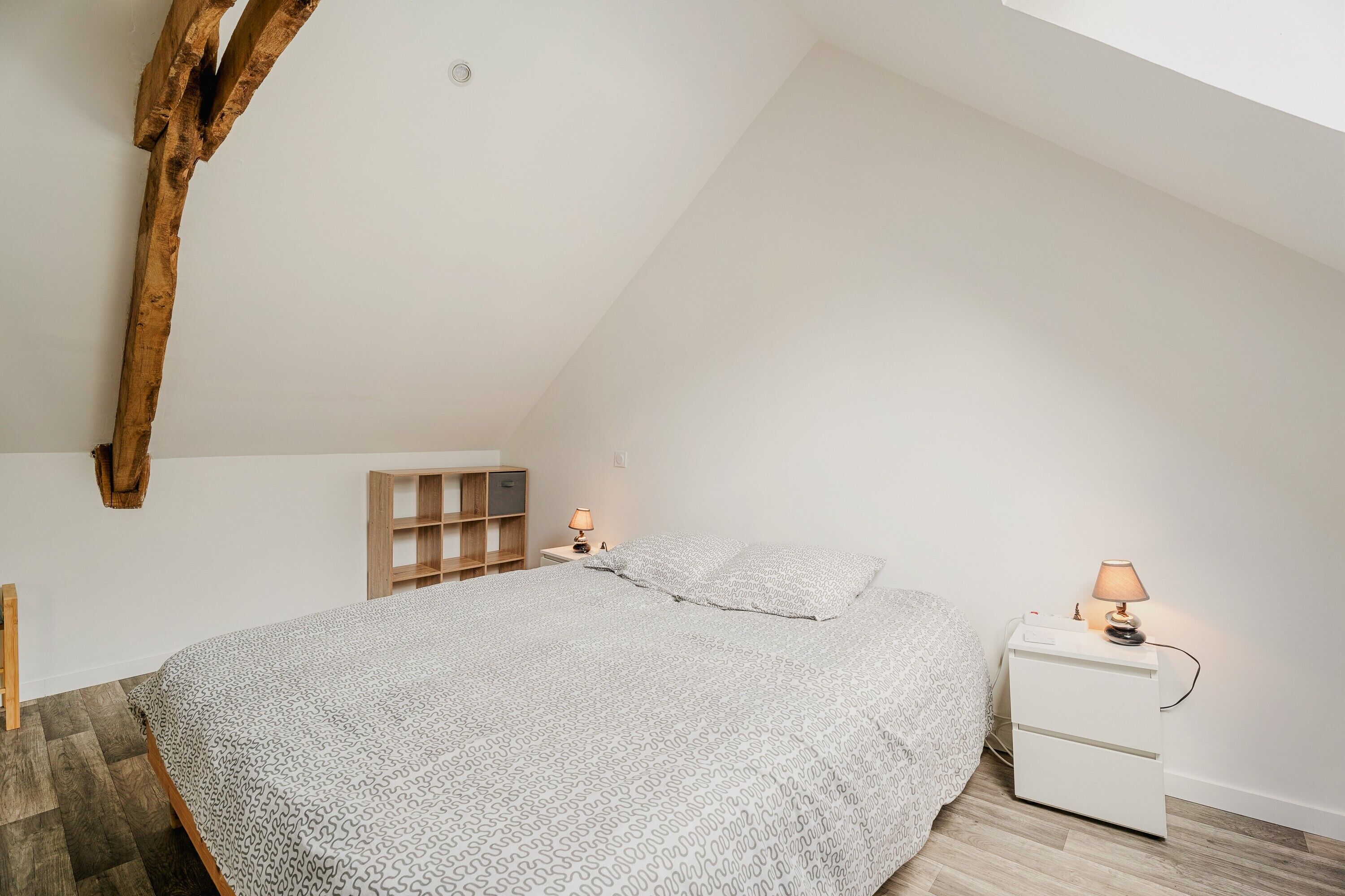 Ein Schlafzimmer mit schrägverdeckter Decke, einem Bett mit gemusterten Betttextilien, einem Nachttisch mit Lampe und einer Holzregaleinrichtung.