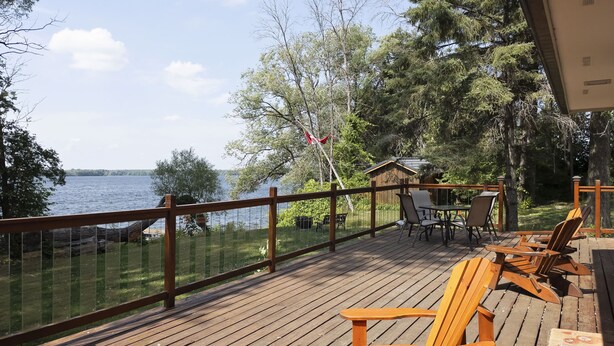 Eine Holzterrasse mit Stühlen und Tisch, mit Blick auf einen See, umgeben von Bäumen und einer Holzhütte.