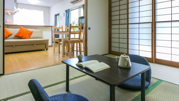 Una habitación tradicional japonesa con suelo de tatami, una mesa baja con un hervidor y un sofá con cojines naranjas.