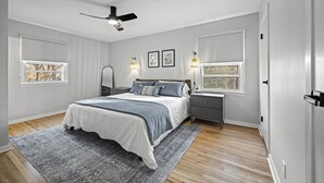 Un dormitorio con cama, mesitas de noche, ventilador de techo y dos ventanas con cortinas.