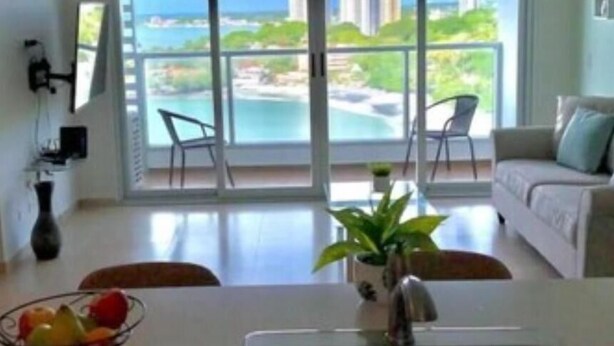 Una sala de estar moderna con un amplio ventanal corredero que da a un balcón con vista al mar y edificios.