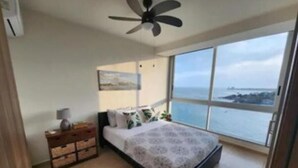 Un dormitorio con cama, una mesita de noche, un ventilador de techo y un ventanal con vistas al mar.