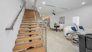 Un intérieur moderne avec un escalier en bois, un coin repas avec un canapé blanc et des coussins bleus, et un fauteuil en cuir noir.