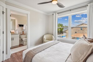 Un dormitorio con una cama grande, ventilador de techo, vistas al exterior y un baño con inodoro y lavamanos.