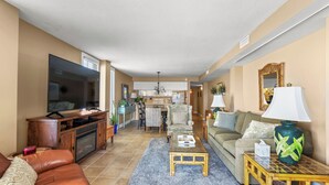 A spacious living room with a leather sofa, a coffee table, and a television. A kitchen is visible in the background with a dining area. - Sea Marsh I 104 - 3BD/2.5BA - Oceanfront (North Myrtle Beach)