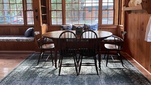 A wooden dining area with a round table and chairs, large windows with a view of the outside, and a hanging light fixture. - Charming, quiet 3-BR cottage; private access to cove and forest trails (Phippsburg)