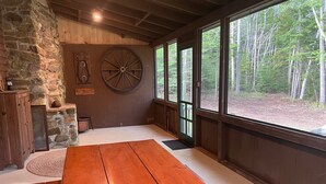 A rustic interior with a stone wall, wooden beams, a large wooden table, and a view of a forest through large windows. - Charming, quiet 3-BR cottage; private access to cove and forest trails (Phippsburg)