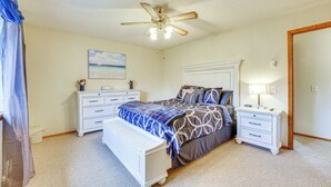 Une chambre à coucher comprenant un lit, des tables de chevet, une commode, un banc, un ventilateur de plafond, un tableau et un miroir.