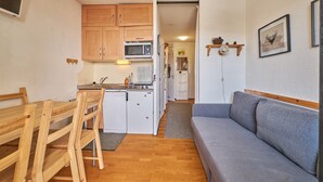 Une cuisine compacte avec des meubles en bois, un four à micro-ondes et un réfrigérateur. On y trouve une table à manger avec des chaises, ainsi qu’un canapé gris dans l’espace salon.